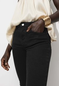 Jean en denim noir au design ajusté, avec des poches avant et une fermeture à bouton argenté. Assorti à un haut léger et fluide et des bracelets en or.