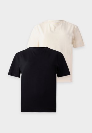 Femme aux longs cheveux portant des lunettes de soleil noires, un t-shirt noir, un pantalon large noir et des baskets noires, debout sur un fond blanc.