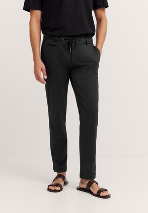 Schwarze Hose mit einem Kordelzugbund, schmaler Passform und Fronttaschen. Getragen mit braunen Sandalen, die eine glatte Stoffstruktur zeigen.