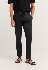 Mann in schwarzen Jogginghosen, schwarzem kurzärmligen Shirt und braunen Sandalen mit zwei Riemen, der mit den Händen in den Taschen steht.