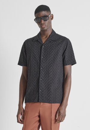 Man draagt een zwart patroon overhemd met korte mouwen en knopen, bruine broek en zwarte vierkante zonnebril tegen een effen achtergrond.