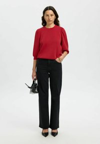 Donna in piedi che indossa un top rosso con maniche a palloncino, jeans neri a vita alta, tacchi appuntiti neri e tiene in mano una piccola borsa nera.