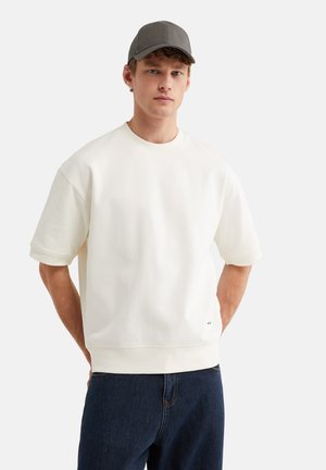 Jonge man met een donkergrijze pet, oversized wit kort mouw sweatshirt en donkerblauwe jeans, staand met handen achter de rug.