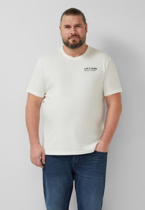 Mann mit Bart, der ein weißes T-Shirt mit kurzen Ärmeln und dem Schriftzug "Let's Unite" sowie eine blaue Jeans trägt, steht vor einem einfachen hellgrauen Hintergrund.