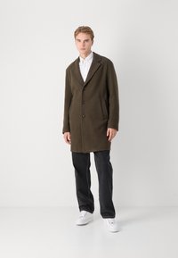 Olivgrön yllekappa med urklippt krage, tre knappar och sidofickor, kombinerad med svarta byxor och vita sneakers.