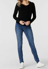 Černý top s dlouhým rukávem a knoflíky, kombinovaný s modrými skinny džínami a bílými teniskami. Oblečení má přiléhavý střih a hladkou strukturu.