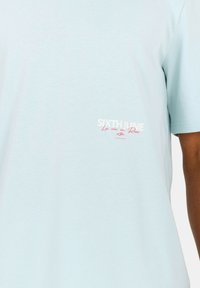 Ljust blå bomullströja med korta ärmar, med vit och rosa tryckt text på bröstet. Rund halsringning, slät yta.