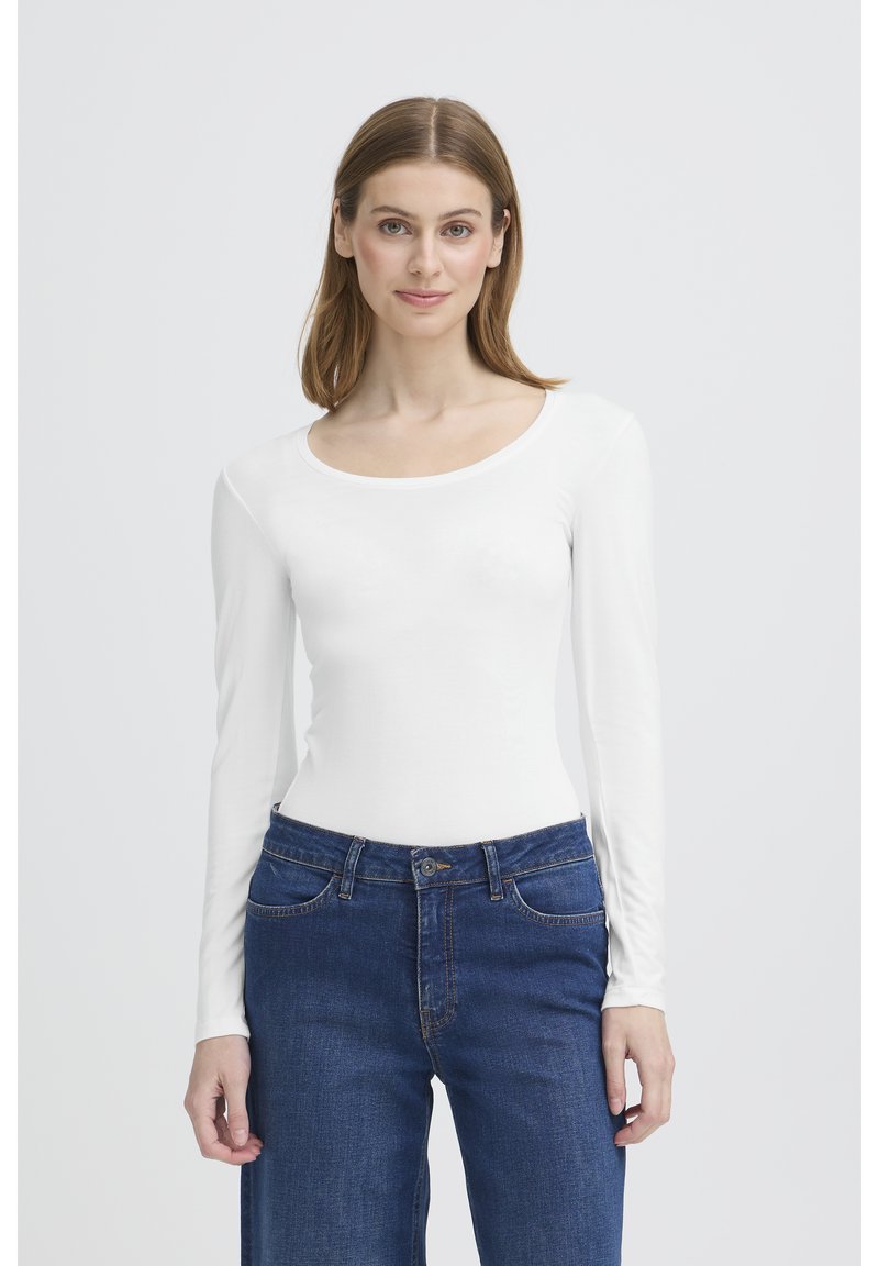 Femme aux cheveux châtain clair portant un haut blanc à manches longues et un jean bleu, debout devant un fond gris clair uni.