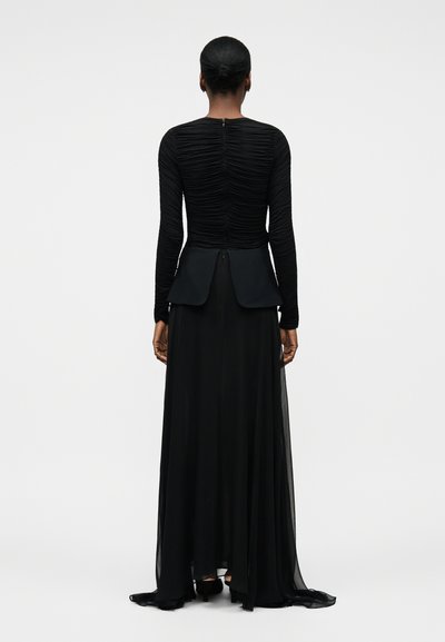 Kvinde med kort hår iført en lang sort ruched top og en flydende sort nederdel, set bagfra mod en ensartet lys baggrund.