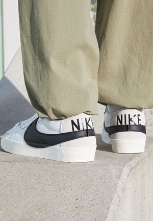 Person trägt weiße Nike-Sneaker mit schwarzem Swoosh und olivgrüner Hose und steht draußen auf einer Betonkante.