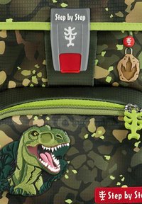 Sac à dos camouflage avec des accents verts, un design de dinosaure en caoutchouc et un fermoir gris portant l'inscription "Étape par Étape." Comprend des détails uniques sur les tirettes de zip.