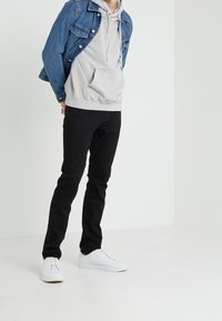 Chaqueta de mezclilla con botones, sudadera gris con bolsillo frontal, pantalones negros ajustados y zapatillas blancas, en un fondo neutro.