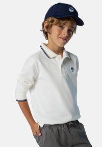 Camisa polo blanca de manga larga con detalles en marino y un logo en el pecho, combinada con pantalones cortos grises de cintura elástica. Una gorra azul marino completa el look.