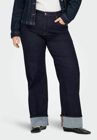 Temno modre kavbojke širokih hlačnic z rdečim kontrastnim šivom, obrobljene zanje z svetlo modrim tkanjem in sprednjo zaponko na gumbe.