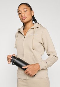 Μπεζ φούτερ με φερμουάρ και κορδόνια, με κεντημένο λογότυπο, συνοδευόμενο από μαύρο μονωμένο παγούρι νερού, κρατημένο σε χέρι.