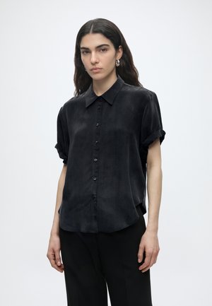 Jeune femme aux longs cheveux foncés porte une chemise noire ample à manches courtes boutonnée et un pantalon noir, debout devant un fond uni.
