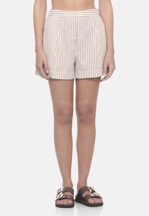 Donna che indossa pantaloncini a righe verticali beige e bianchi e sandali marroni con grandi fibbie dorate, in piedi davanti a uno sfondo bianco.