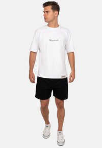 Witte katoenen t-shirt met korte mouwen en een ronde hals, voorzien van zwarte cursieve letters. Gecombineerd met zwarte shorts en witte sneakers.