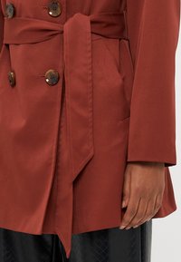 Pasasti rjave barve s pasom in velikimi rjavimi gumbi, sprednjim žepom ter črne teksturirane hlače z rahlo ohlapnimi rokavi in manšetami.