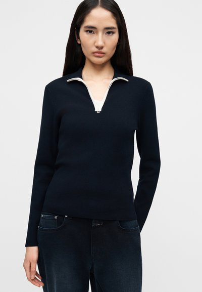 Femme portant un pull-over bleu marine à manches longues avec un col zippé blanc et un jean foncé, debout devant un fond clair uni.