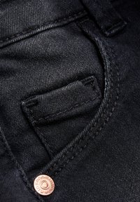 Svart denimstoff med en liten, horisontal welt-lomme. Har kobberdetaljer og fin søm langs kantene. Glatt tekstur.