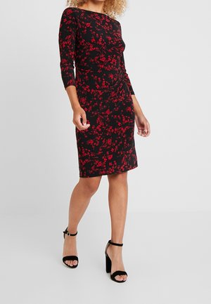 Femme portant une robe noire jusqu'aux genoux avec des motifs floraux rouges et des manches trois-quarts, assortie à des sandales noires à talons hauts avec bride à la cheville.