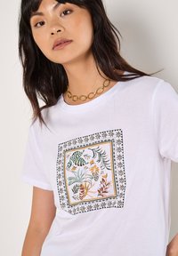 Maglietta bianca in cotone con una stampa botanica colorata incorniciata in nero e oro, con scollatura tonda e maniche corte.