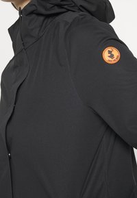 Czarna kurtka z kapturem o smukłym kroju, gładkiej fakturze i pomarańczowej, okrągłej naszywce z logo na lewym ramieniu.