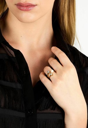 Donna dalla pelle chiara che indossa una camicetta nera trasparente mostrando un anello d'oro con due diamanti quadrati e una perla sul dito.