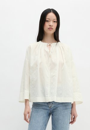 Frau mit langen schwarzen Haaren trägt eine lockere, cremeweiße Bluse mit Bindeband vorne und hellblaue Jeans vor weißem, einfarbigem Hintergrund.