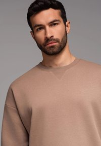 Beige Crewneck-Sweatshirt mit strukturierter Oberfläche, versehen mit einer V-förmigen Nahtdetails am Kragen und übergroßen Ärmeln.