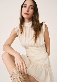 Joven con cabello largo y castaño lleva un vestido sin mangas crema plisado y botas con estampado de piel de serpiente, sentada con una pierna levantada.