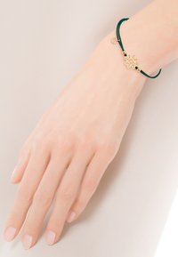 Zielona bransoletka z cordu z centralnym złotym emblematem w kształcie kwiatu i złotymi koralikami, zamykana na prosty zamek. Gładka faktura.