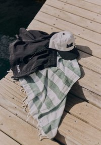 Shorts noirs, casquette blanche et noire, et serviette rayée vert et blanc posés sur un ponton en bois au bord de l'eau.