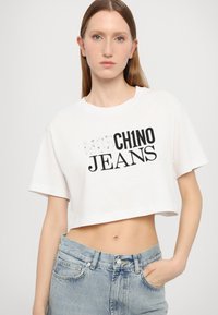 Balta trumpa marškinėliai su juodu tekstu "MOC CH1NO JEANS", trumpomis rankovėmis ir laisvu siluetu, derinami su šviesiai mėlynomis aukštu juosmeniu džinsais.