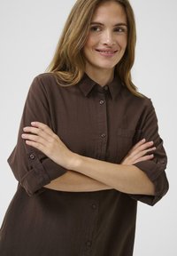 Barna, gombos ing puha anyagból, gallérral, mellzsebbel és felhajtott ujjal, melyeken gomb díszítés található.