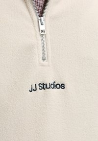 Beige fleece quarter-zip pullover with a silver zipper and embroidered "JJ Studios" in black. Features a soft texture and relaxed fit.