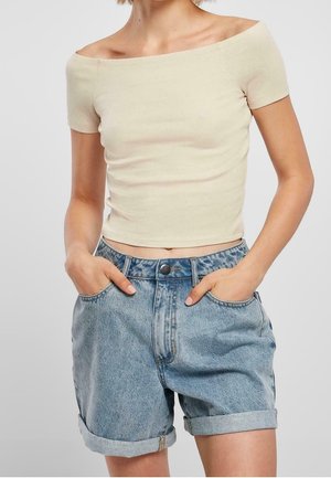 Kobieta ubrana w beżowy, odkrywający ramiona krótki top i jasnoniebieskie podwinięte szorty jeansowe, z rękami w kieszeniach na jednolitym tle.