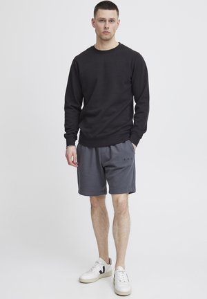 Schwarzer Pullover mit Rundhalsausschnitt, kombiniert mit grauen Shorts, die ein kleines "A.R.C."-Logo tragen. Weiße Sneaker mit schwarzen Akzenten runden den Look ab.