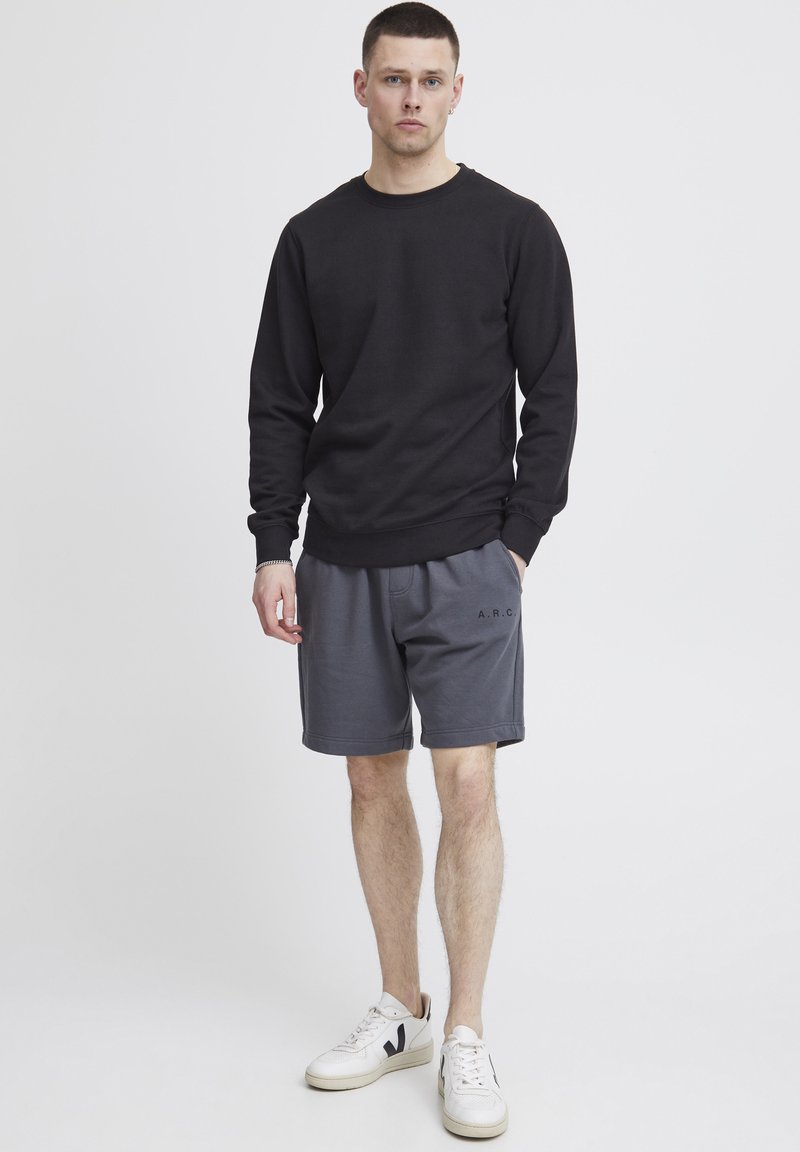 Felpa nera con scollatura a girocollo, abbinata a pantaloni corti grigi con un piccolo logo "A.R.C.". Sneakers bianche con dettagli neri completano il look.