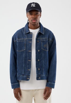 Noor mees kannab tumel sinist teksajope valge särgi, beežide pükste ja tumel sinise New York Yankeesi pesapalli nokamütsiga.