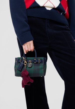 Une personne portant un pull bleu marine et un pantalon foncé tient un petit sac à main vert à motif tartan, avec des garnitures dorées et un charm bordeaux en forme de chien.