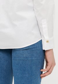 Hvit skjorte med glat tekstur og gullknappdetail på mansjetten, parret med tettsittende blå denimbukser.