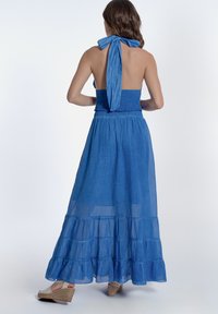 Lang blauwe jurk met een smock-bodice, open rug en halterstrik. Heeft een gelaagd rokdesign en lichte stof, wat zorgt voor een vloeiende uitstraling.