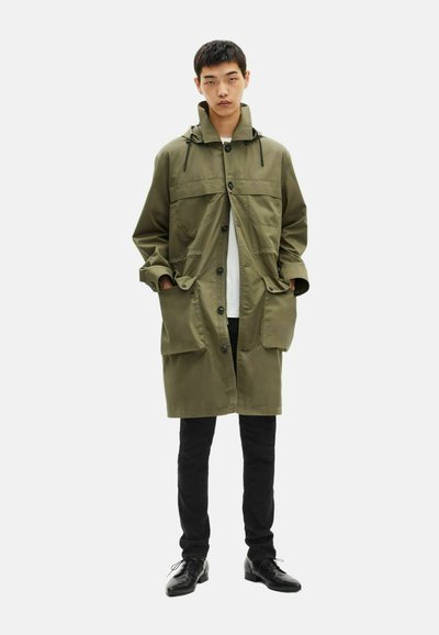 Giovane uomo che indossa un lungo cappotto verde oliva con bottoni e grandi tasche, pantaloni neri e scarpe nere con lacci, in piedi con le mani in tasca.