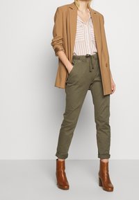 Bruine blazer, gestreept lichtgekleurde overhemd, olijfgroene tapered broek en bruine enkellaarzen. Heeft een relaxte pasvorm en opgerolde mouwen.