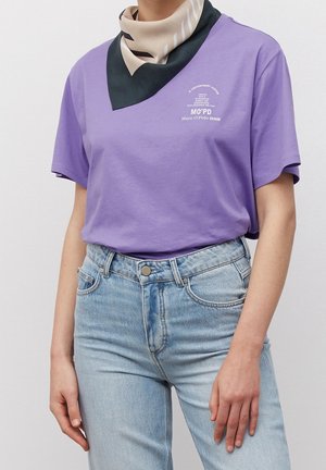 Person trägt ein lila Marc O'Polo Denim T-Shirt, hellblaue, hoch taillierte Jeans und einen beige-schwarz gemusterten Schal.