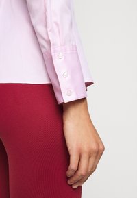 Mano appoggiata di lato, indossa una camicia a maniche lunghe rosa con tre bottoni sui polsini e pantaloni rossi su uno sfondo semplice.