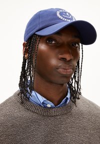 Navyblaues Baseballcap mit einem weißen Logo, getragen über einem hellblauen gestreiften Hemd und einem strukturierten grauen Pullover. Lange, gedrehte Haare sind sichtbar.