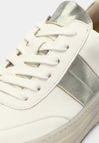 White leather sneaker featuring a metallic silver accent on the heel and a grey lateral stripe, with flat white laces and textured surface.